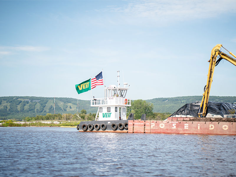 Marine contractors use powerful tugboats and excavator-equipped barges to move heavy loads and handle sediment control during foundation and seawall construction. These vessels support the logistical backbone of large-scale piling and coastal infrastructure projects across waterways. Photo Credit: Veit